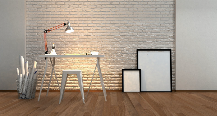 Minimalist workspace with a white desk, modern lamp, stool, and art supplies. Rolled papers in a metal bin sit to the left, with two empty picture frames leaning against a white brick wall on a wooden floor.
