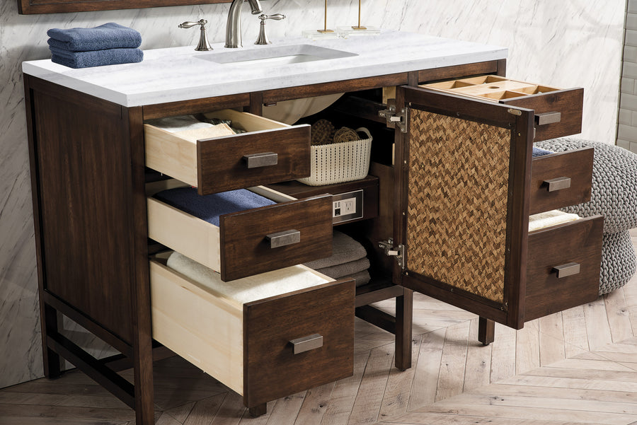 Dark wood bathroom vanity with white countertop and sink, open drawers revealing neatly folded towels and organized storage baskets, against a light herringbone-patterned floor and marble wall.