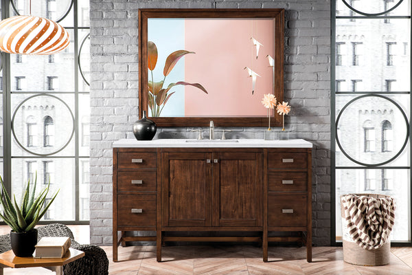 A modern bathroom with a wooden vanity, white countertop, and sink, set against a gray brick wall. A large framed art piece, plants, and decorative accents are arranged on and around the vanity. Tall windows let in natural light.