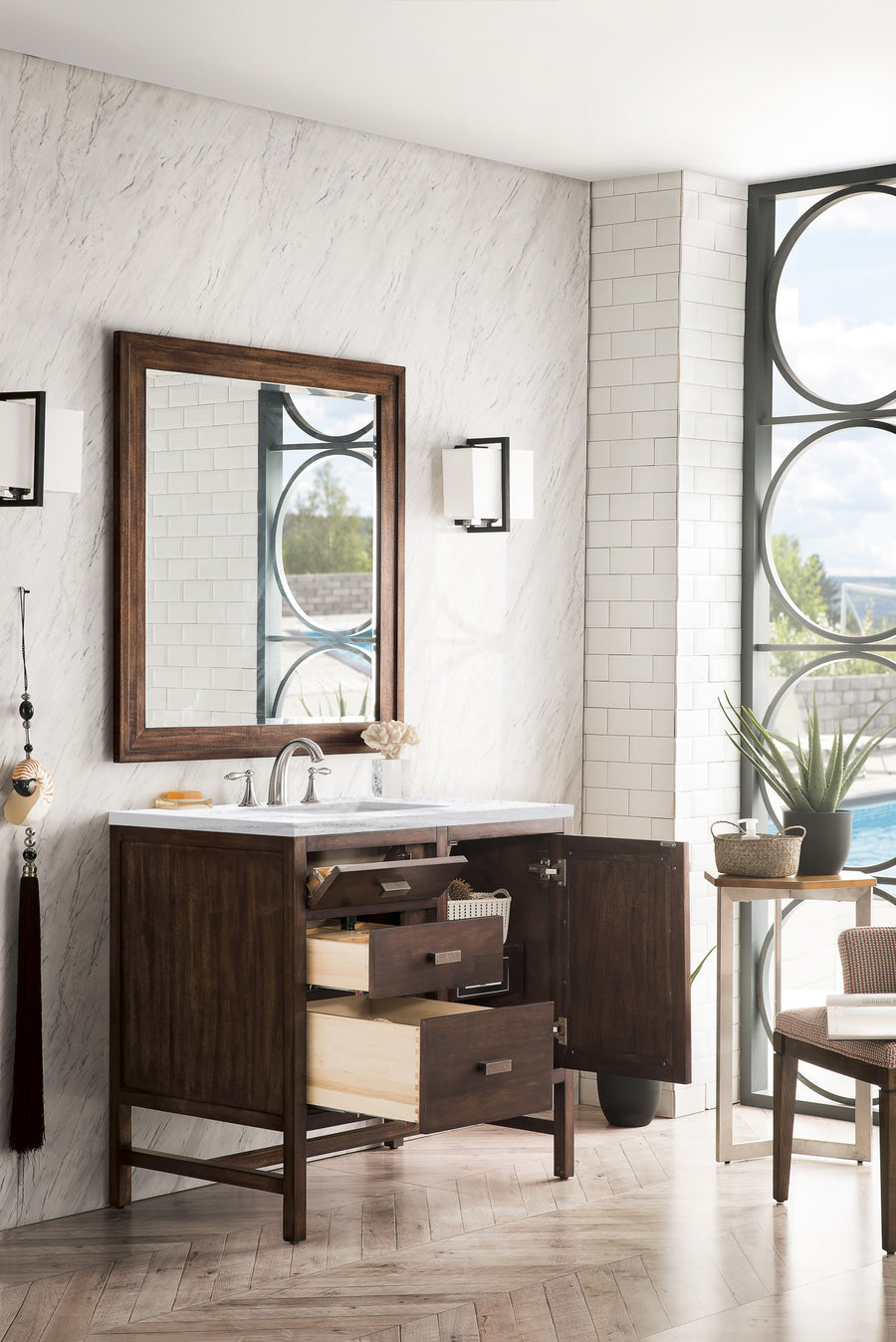 A modern bathroom with a dark wood vanity, white marble countertop, and open drawers. A large mirror is above the sink, flanked by wall lights. There are plants, a basket, and a large window with a pool view.