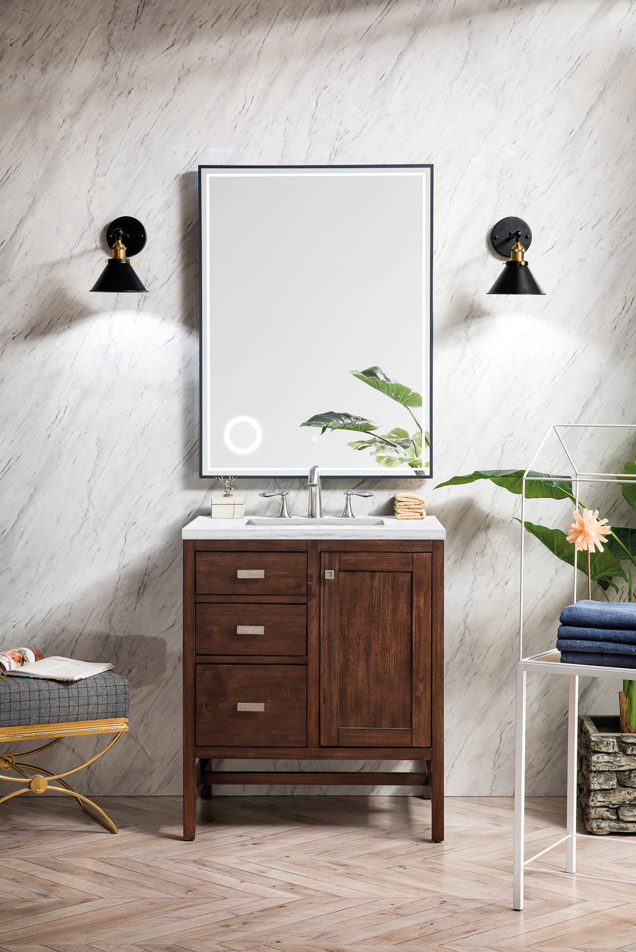 A modern bathroom with a dark wood vanity, white countertop, large rectangular mirror, two black wall sconces, and potted plants on either side, set against a light marble-patterned wall.