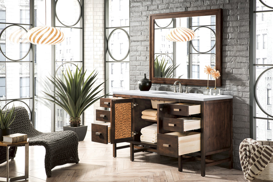 Modern bathroom with large windows, a wooden vanity with open drawers revealing towels, a mirror, two pendant lights, a plant, and a woven chair on a herringbone-patterned wood floor.