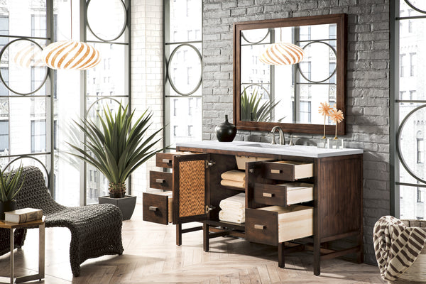 Modern bathroom with large windows, a wooden vanity with open drawers revealing towels, a mirror, two pendant lights, a plant, and a woven chair on a herringbone-patterned wood floor.