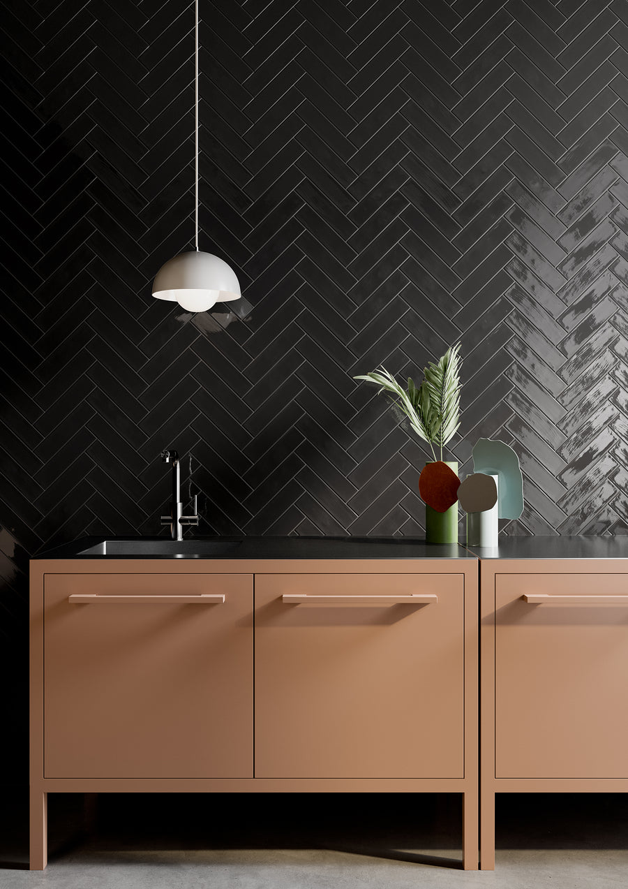 Minimalist kitchen with matte pink cabinets, a sleek black countertop, and a modern black herringbone tile backsplash. A pendant light hangs above, and vases with green leaves and decor sit near the sink.
