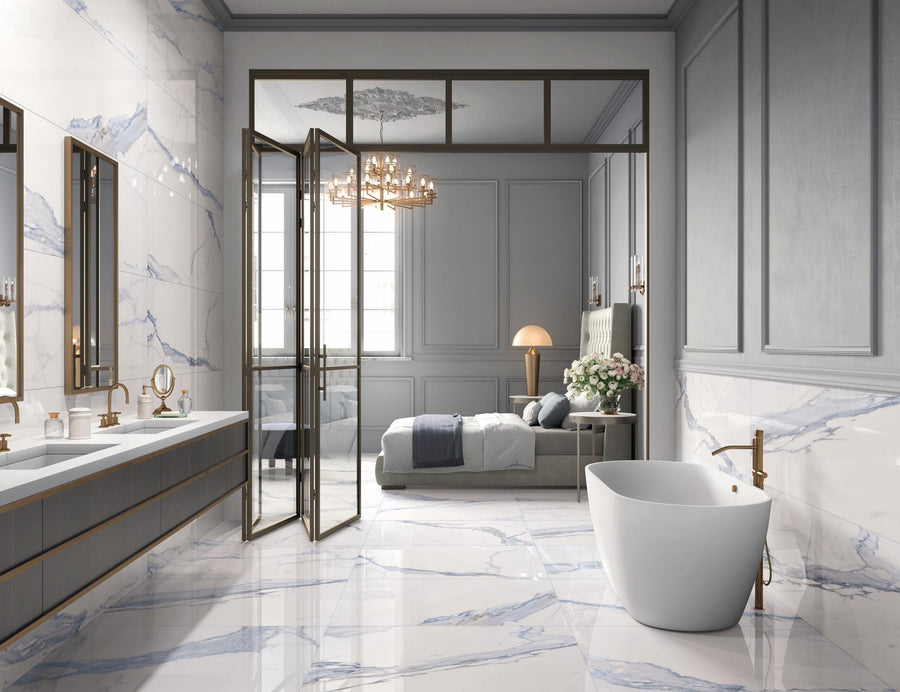 A luxurious bathroom with marble floors and walls, double sinks, a freestanding bathtub, and brass fixtures opens into a stylish bedroom with an upholstered bed, lamp, and chandelier.