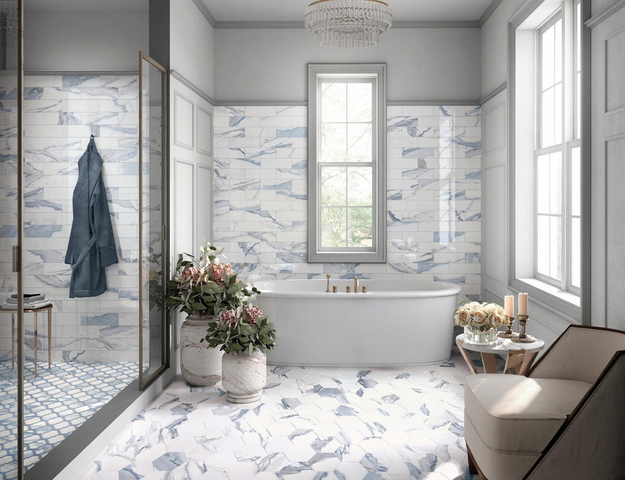 Bright bathroom with a freestanding tub, marble-patterned tiles on walls and floor, a window above the tub, two potted flower arrangements, gold fixtures, and a beige armchair with candles on a side table.