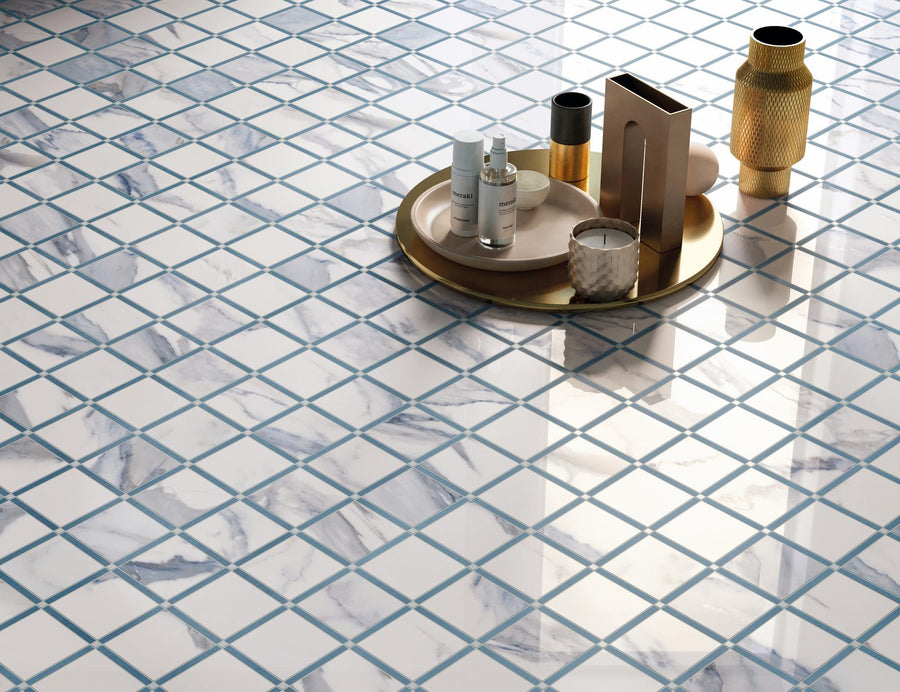 A brass tray with skincare bottles and decorative items sits on a floor of white and gray marble tiles with blue grout arranged in a grid pattern.