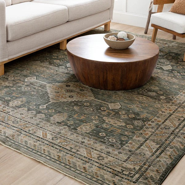 A round wooden coffee table sits on a patterned area rug in a living room with a light-colored sofa and a wooden chair, creating a cozy and stylish space with neutral tones.