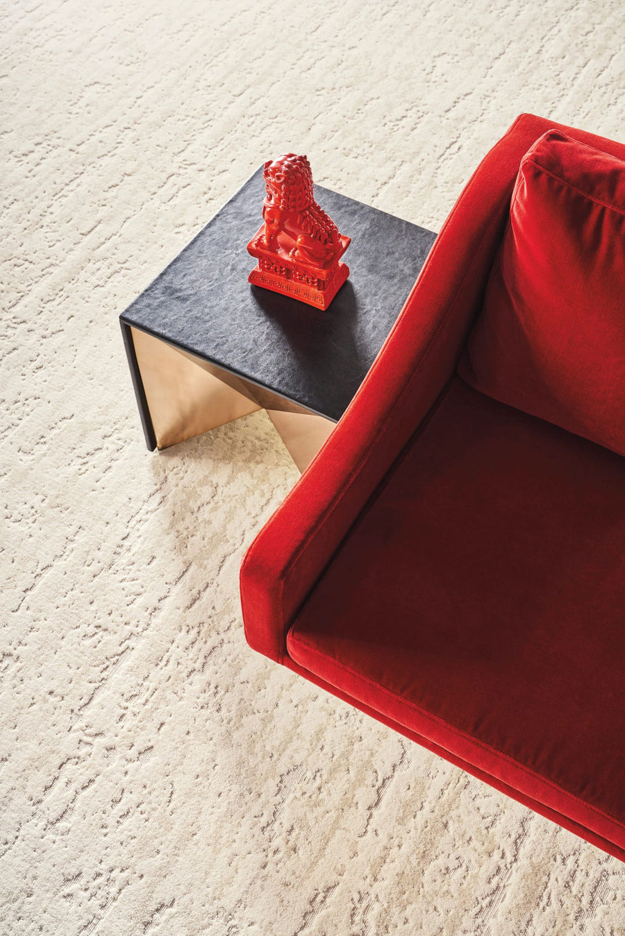 A modern scene with a red velvet sofa next to a black and gold side table, topped with a small red decorative lion statue, all set on a textured cream carpet.