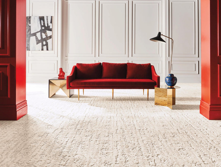 A modern living room with a red velvet sofa, gold side tables, a blue vase, and a black floor lamp. The space features white paneled walls, bold red columns, abstract artwork, and a light textured carpet.