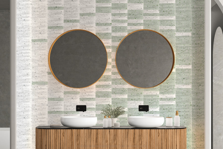 A modern bathroom with two round mirrors above a wooden vanity, featuring vessel sinks and black faucets. The wall has a mix of light green and white tiles, and toiletries are neatly arranged beside a small plant.