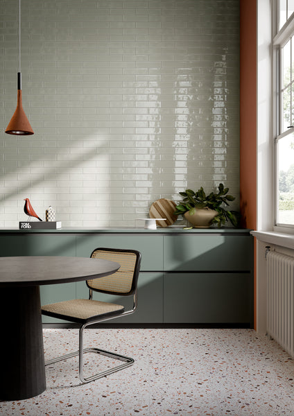 Modern kitchen with glossy light gray subway tiles, green cabinets, a round dark table, a cane chair, potted plant, decorative objects, terrazzo floor, and large window letting in natural light.