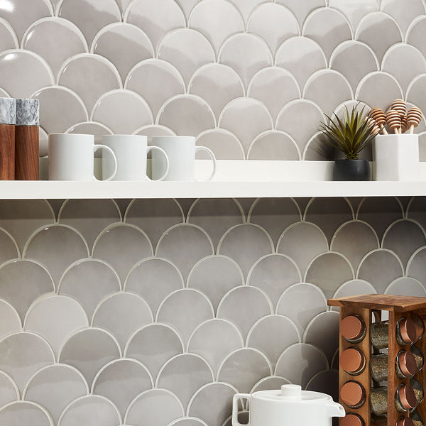 A kitchen with a scalloped, fish scale-patterned beige tile backsplash, white shelves holding white mugs, small containers, a potted plant, and a wooden wine rack below.