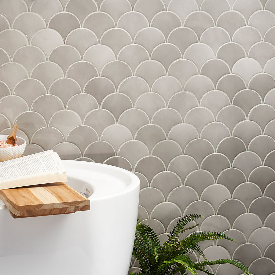 A modern bathroom features gray fish scale tile walls, a white bathtub with a wooden bath tray holding an open book and a bowl, and a green fern plant nearby.