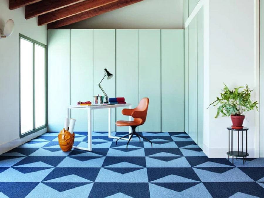 A modern office featuring Commercial Modular Carpet Tiles in geometric blue, light blue walls, a white desk with lamp and books, an orange chair, floor vase, and a potted plant on a black stand—perfect for any commercial space.