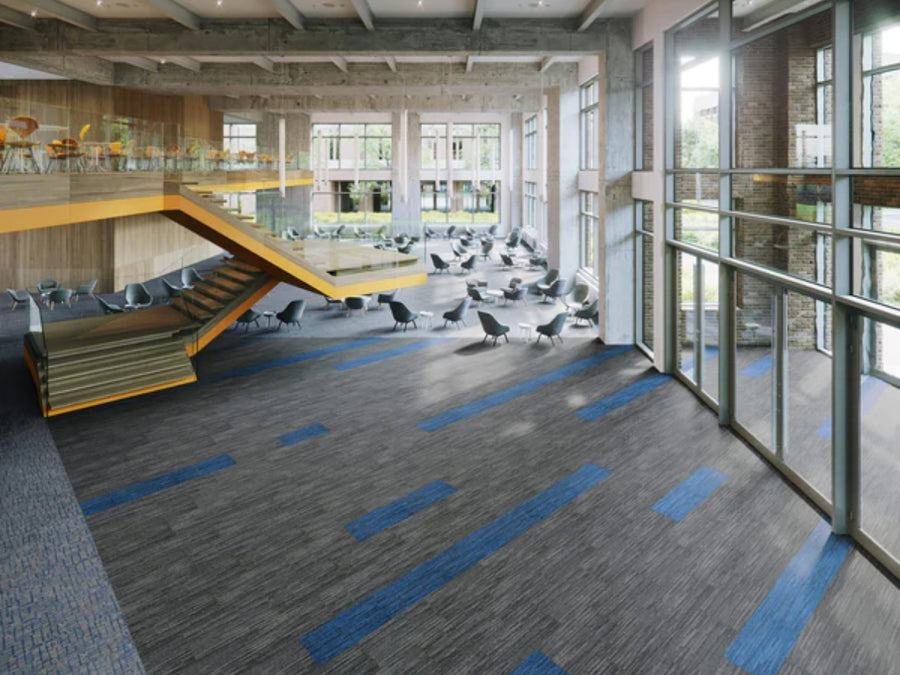 Spacious, modern commercial space featuring large windows, open staircase, gray chairs and tables, and striped Commercial Modular Carpet Tiles in gray and blue tones. Natural light fills the room with trees visible outside.