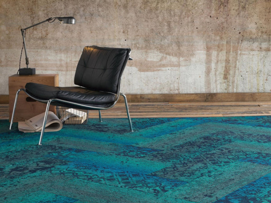 A modern black leather chair and small wooden table with a desk lamp rest on Commercial Modular Carpet Tiles in blue, set against a textured wall—an ideal choice for stylish commercial spaces.