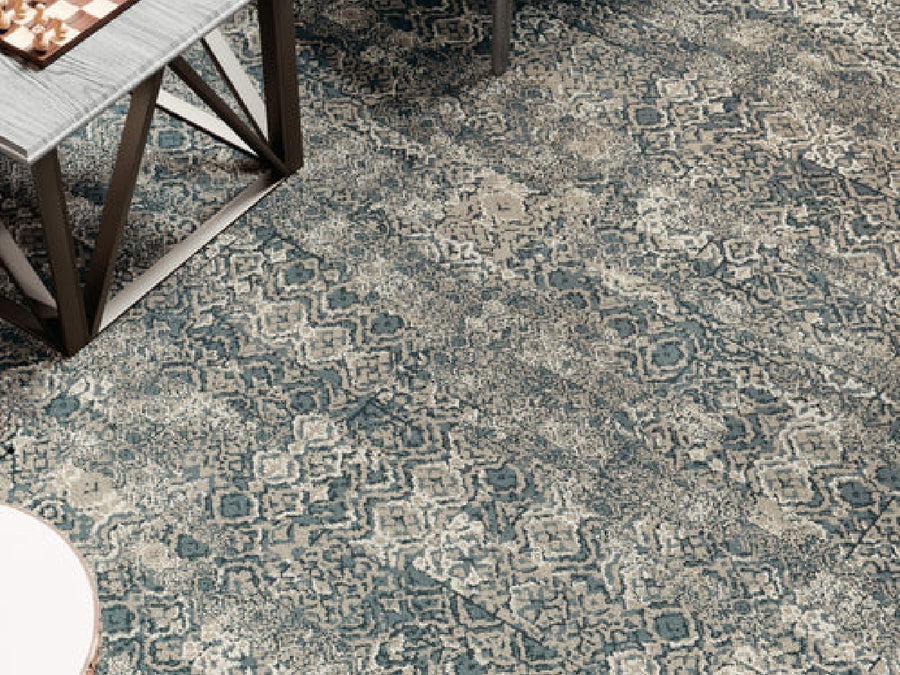 A close-up of Commercial Modular Carpet Tiles in blue and beige with geometric and abstract patterns, installed in a commercial space next to a light table with metal legs and part of a round table in the lower left corner.