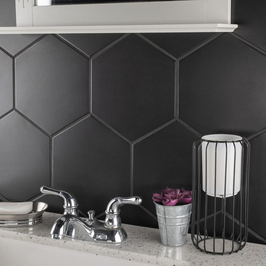 A bathroom countertop with a chrome faucet, a small metal tray, a potted purple flower, and a metal lantern-style holder. The wall features large, matte black hexagonal tiles.