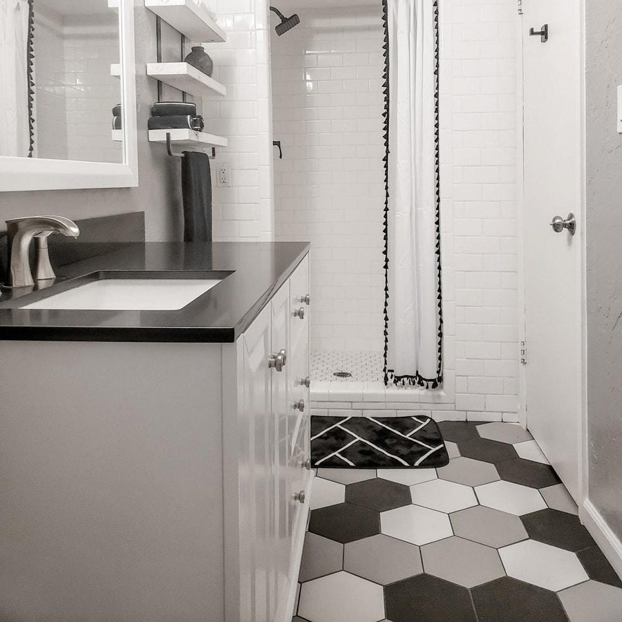 A modern bathroom with a black and white hexagonal tile floor, a white vanity with a black countertop, a sink, a wall mirror, shelves, and a walk-in shower with white subway tiles and a shower curtain.