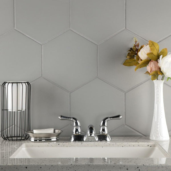A modern bathroom sink with a chrome faucet, gray hexagon tile backsplash, a white rectangular vase with flowers, a wire holder with rolled towels, and a soap dish on a speckled countertop.
