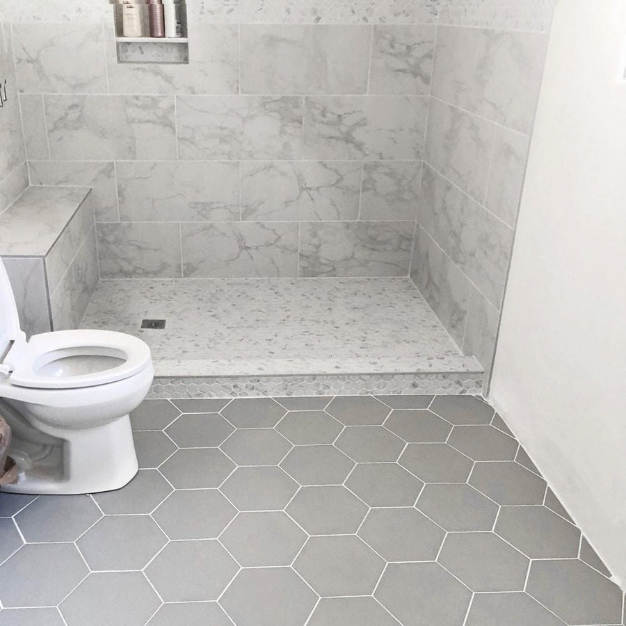 A bathroom with a toilet on the left, gray hexagonal floor tiles, and a walk-in shower featuring light marbled wall tiles and a matching shower curb. Toiletries are visible on a ledge inside the shower.