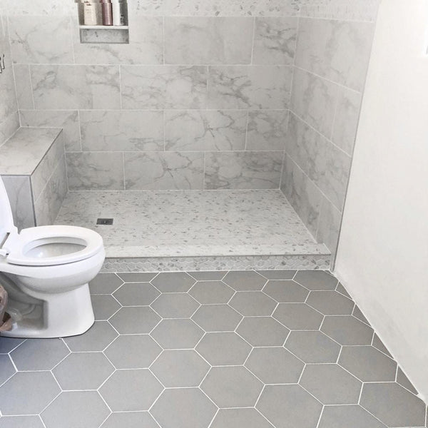 A bathroom with a toilet on the left, gray hexagonal floor tiles, and a walk-in shower featuring light marbled wall tiles and a matching shower curb. Toiletries are visible on a ledge inside the shower.