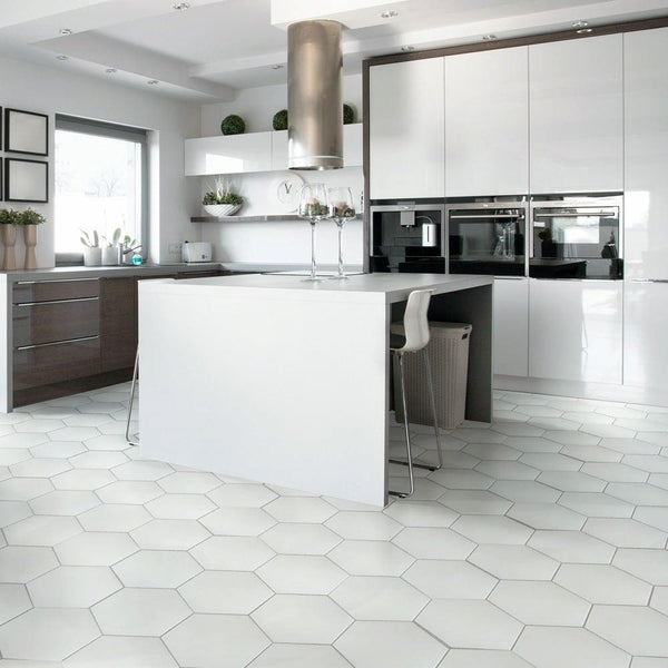 Modern kitchen with white hexagonal tile flooring, glossy white cabinets, a central island with two stools, built-in appliances, and minimalist decor. Large window provides natural light.