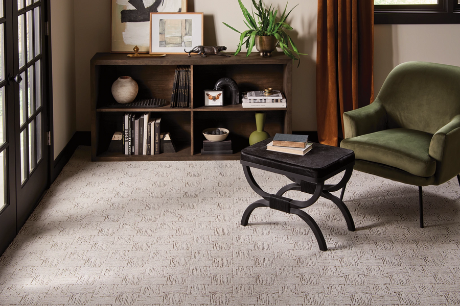 A modern living room with a green armchair, a black ottoman, a wooden console table with books and decor, a potted plant, and artwork on the wall, all on a light-patterned carpet.