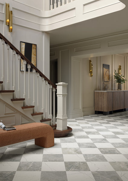 A modern foyer with Monaco 12x12 Glazed Porcelain Tile in a checkered pattern, a wooden staircase with white railings, abstract wall art, a brown cushioned bench, and a wooden console table topped with a flower vase.
