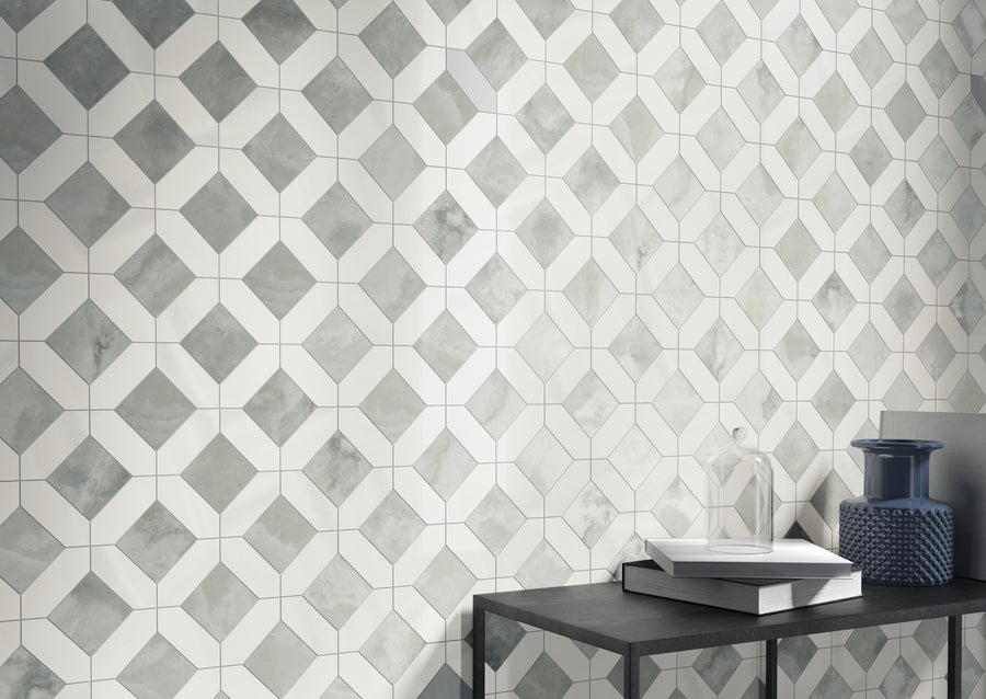 A geometric tiled wall with a pattern of white diamonds and gray squares. In front, a black table holds stacked books, a clear glass decor piece, and a textured blue vase.