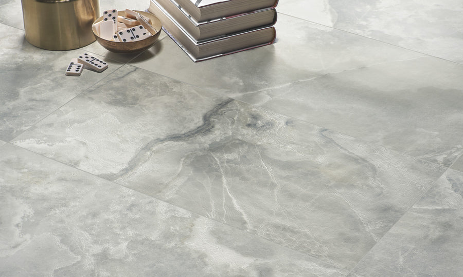 A close-up of gray marble floor tiles with a natural, veined pattern. Nearby are a stack of books, a metallic container, and a small bowl with domino tiles scattered around it.