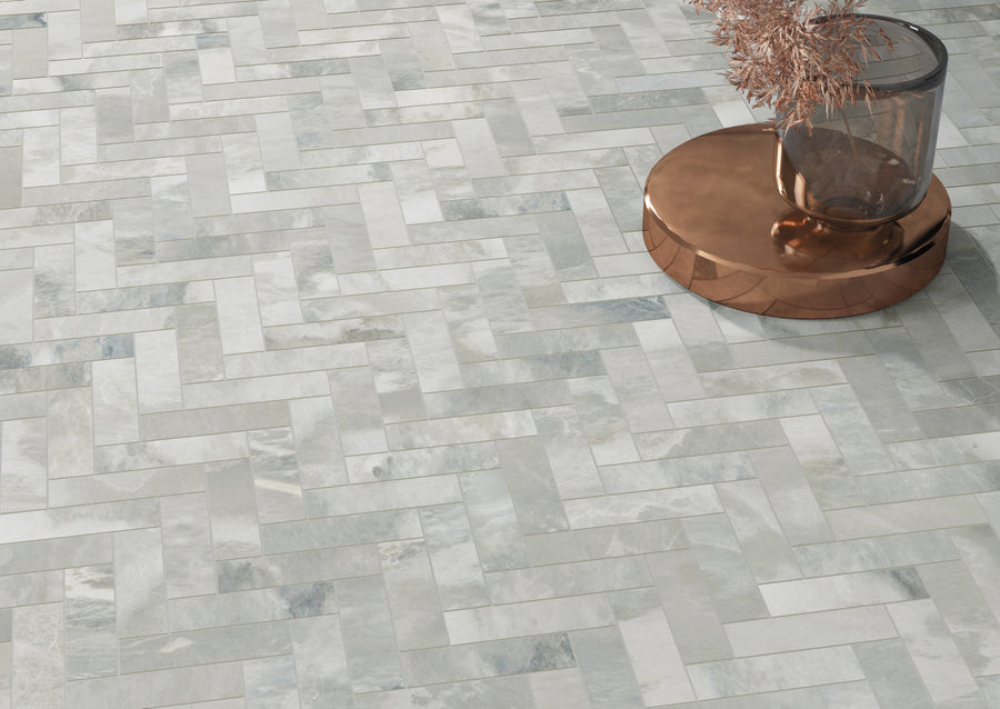 A glass vase with dried branches sits on a round, copper-colored tray placed on a floor covered with light gray and white herringbone-patterned tiles.