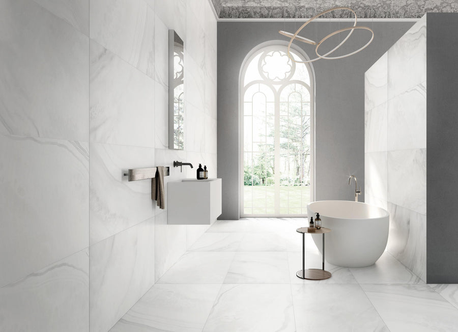 Modern bathroom with marble walls and floors, a freestanding oval bathtub, minimalist sink with mirror, towel rack, round side table, and a large arched window letting in natural light. Contemporary chandelier overhead.
