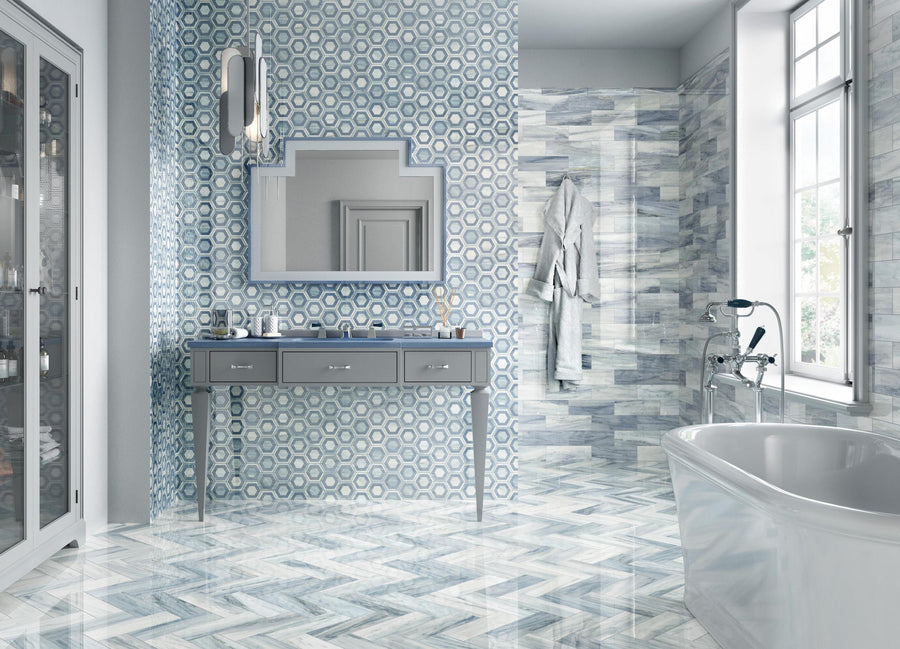 Modern bathroom with blue and white geometric tiles, a large mirror above a blue vanity, glass shower with towels and robes, and a white bathtub near tall windows letting in natural light.