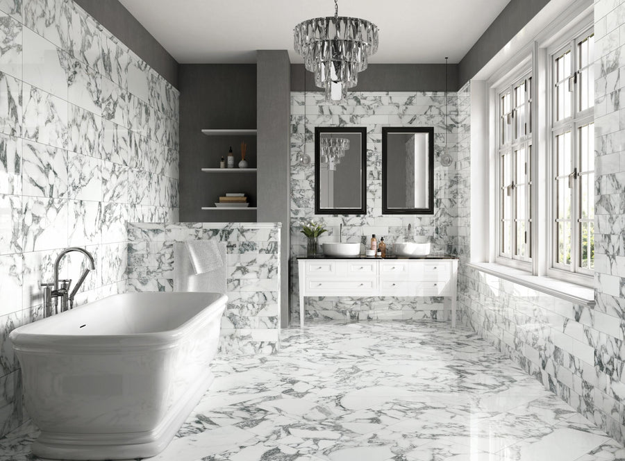 Luxurious bathroom with white marble floors and walls, freestanding bathtub, double sink vanity with two mirrors, shelves, and large windows letting in natural light. Elegant chandelier hangs from the ceiling.