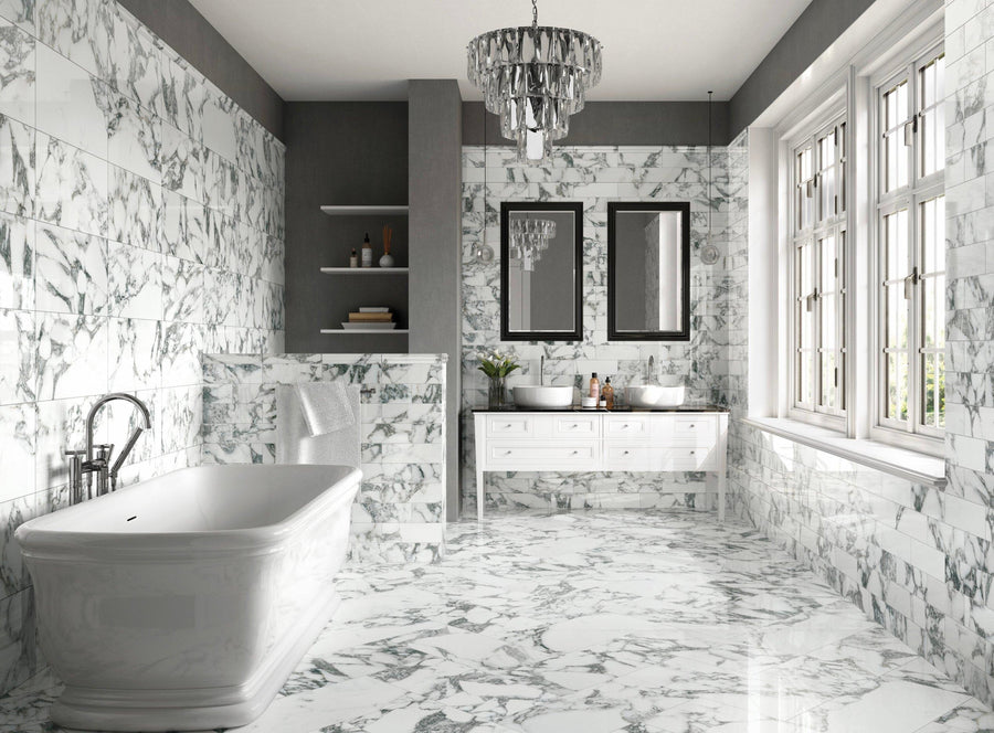 Luxurious bathroom with white and gray marble floors and walls, a freestanding bathtub, double sink vanity with mirrors, shelves with decor, large windows, and a modern chandelier.