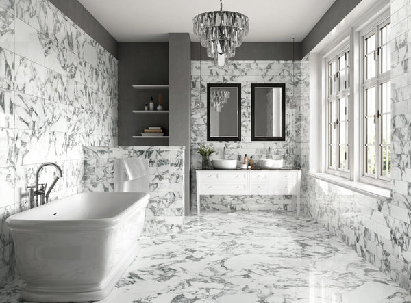 Luxurious bathroom with white marble floors and walls, freestanding bathtub, double sink vanity with two mirrors, shelves, and large windows letting in natural light. Elegant chandelier hangs from the ceiling.