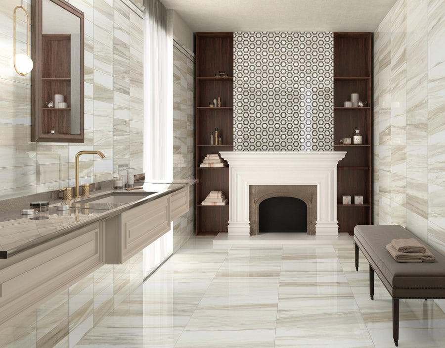 Elegant bathroom with marble floors and walls, double sink with gold faucets, large mirror, fireplace with patterned tile accent, built-in shelves, and a cushioned bench with towels. Natural light from a sheer curtained window.
