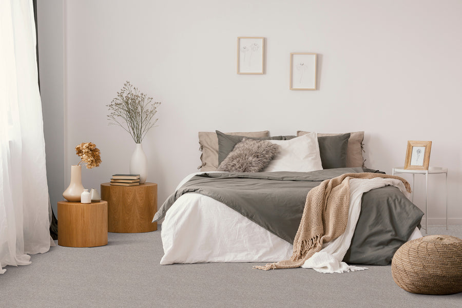 A cozy, minimalist bedroom with a neatly made bed in gray and white bedding, a beige throw blanket, light wood side tables with vases and books, wall art, a pouf, and soft natural light coming through a window.