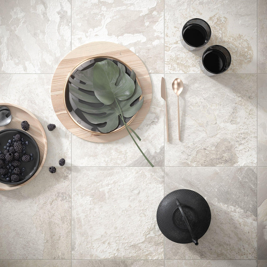 A top view of a stylish setting on beige stone tiles, featuring a black teapot, two black cups, a light wood tray with a green leaf, a plate of blackberries, and a gold spoon and knife.
