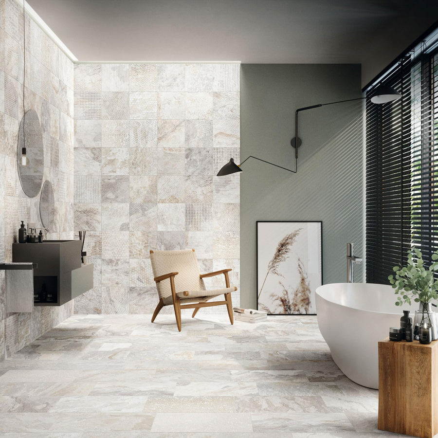 A modern bathroom with stone tile walls and floor, a freestanding white bathtub, a wooden chair, a wall-mounted sink, a round mirror, a black lamp, large window blinds, and a framed botanical print on the floor.