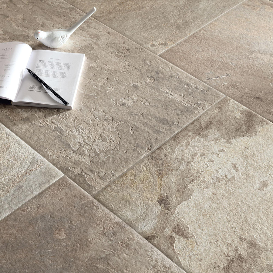 A black pencil and an open book with a small white ceramic bird rest on large, textured, beige stone tiles. The tiles have a natural, uneven surface with subtle variations in color.