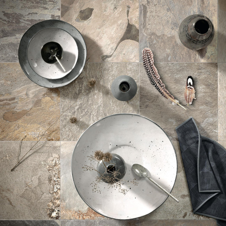 A top-down view of a textured stone tile surface with ceramic bowls, small vases, dried plants, a spoon, feathers, and a folded gray napkin arranged decoratively.