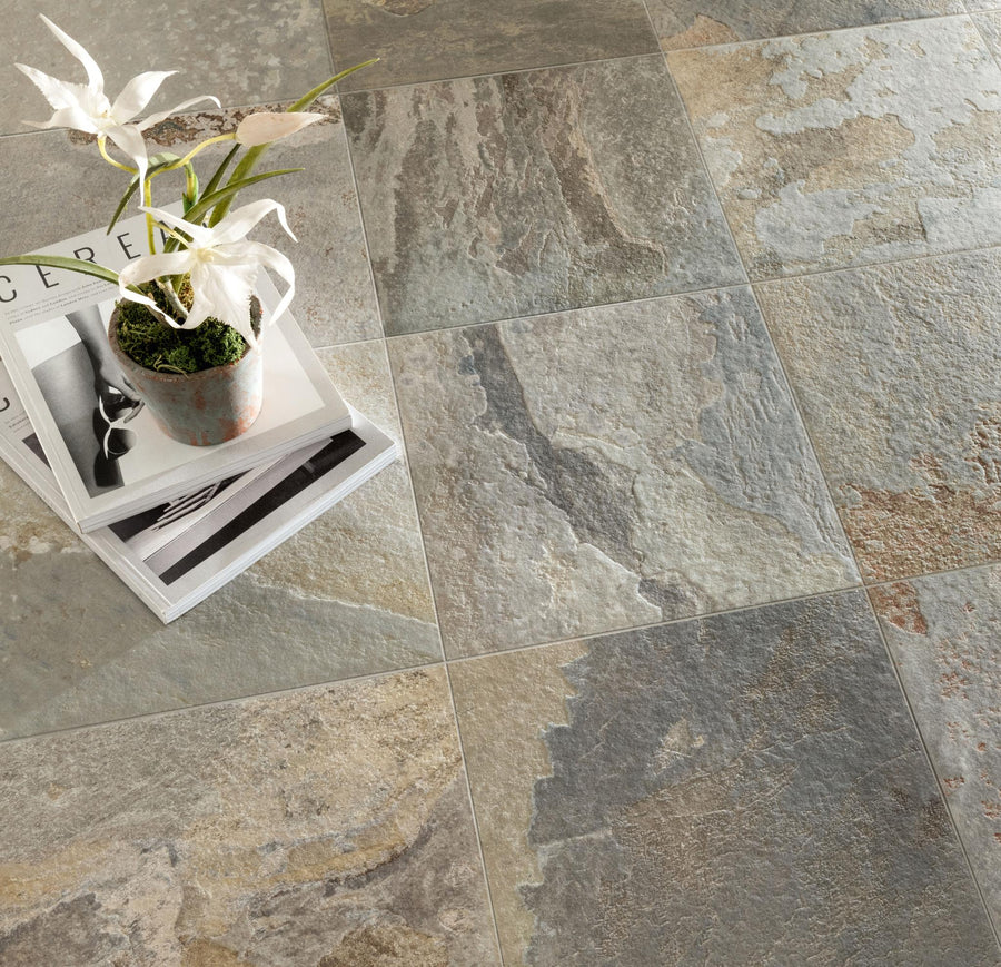 A stack of magazines with a potted plant sits on a textured, multicolored stone tile floor in shades of gray, beige, and light brown.