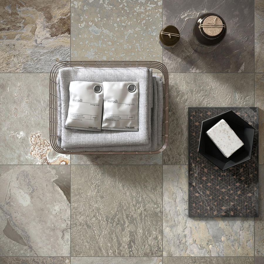 A wire basket with folded gray towels and two white sachets sits on stone tiles. Nearby, there’s a brown bottle, a cup, a patterned tray holding a black dish, and a bar of soap. The scene is viewed from above.