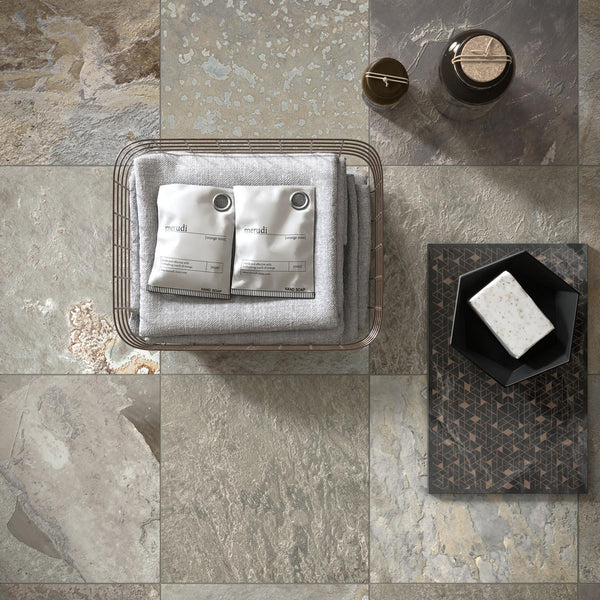 A wire basket with folded gray towels and two white sachets sits on stone tiles. Nearby, there’s a brown bottle, a cup, a patterned tray holding a black dish, and a bar of soap. The scene is viewed from above.