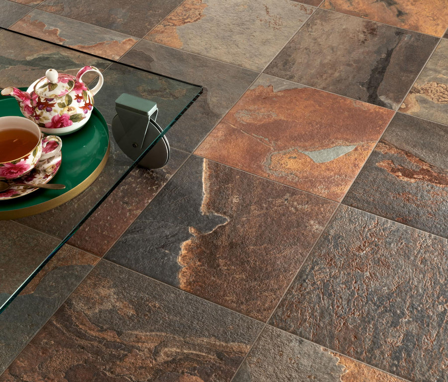 A glass table with a floral teapot, teacup, and a green tray sits atop textured, multicolored stone tile flooring in brown, gray, and rust tones.