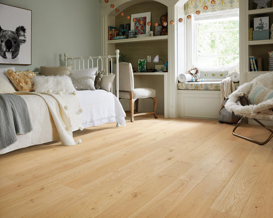 A cozy, sunlit bedroom with light wood flooring, a white metal bed, soft pillows, a built-in desk, shelves with books, and a cushioned window seat overlooking greenery. Warm, inviting, and well-decorated.