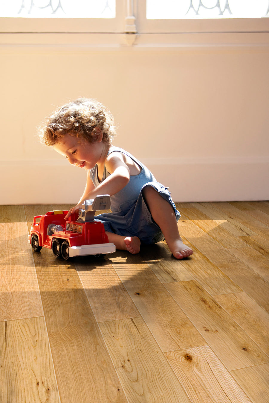 A young child with curly hair sits on a wooden floor in sunlight, playing with a red toy fire truck near a bright window.